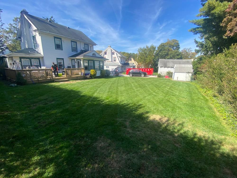 Spacious backyard with green lawn, two houses, and a red truck under a clear blue sky.