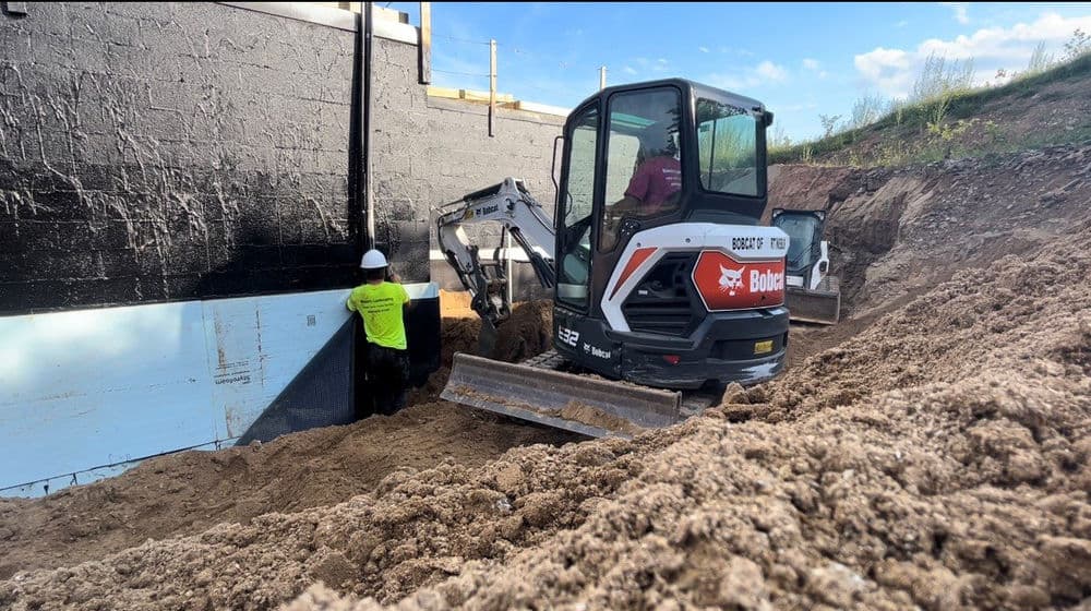 Excavator digging near a basement wall, with a worker in safety gear overseeing the operation.